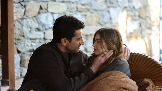 A couple sits close on a wooden bench, gazing at each other while sharing a gentle moment under a brown blanket against a stone wall background