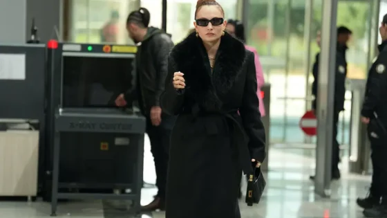 Woman in a black belted coat with a fur collar and dark sunglasses walks through an airport security area, carrying a black handbag.