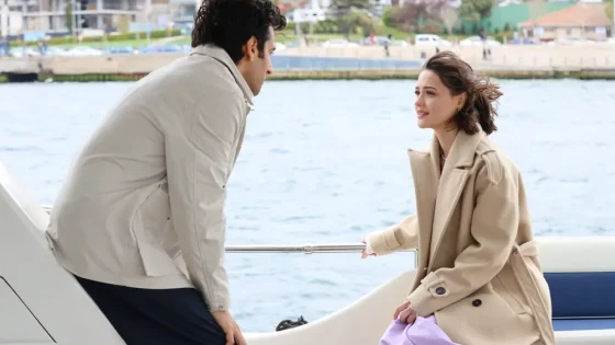 A man and a woman sit and lean toward each other on a yacht by the railing, chatting with a waterfront city in the background.