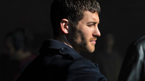 Profile of a man with short dark hair and stubble, wearing a dark coat, looking to the right.