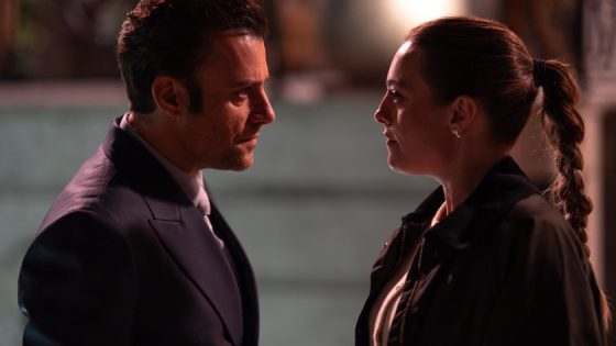 Two people face each other in a dimly lit outdoor setting, engaged in a serious conversation at night.