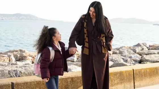 Woman and young girl hold hands and smile as they walk along a rocky seaside promenade by the water.