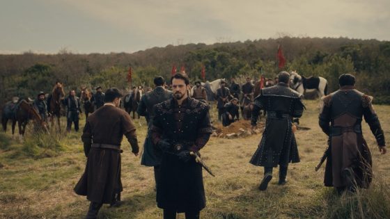 Group of armored medieval soldiers on a grassy field, central figure facing the camera with others and red banners in the background.