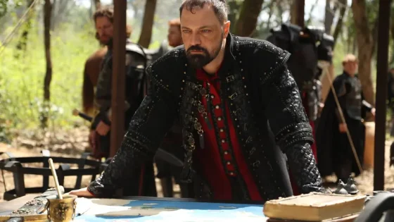 Bearded man in black studded armor studies a map on an outdoor table, with armed companions in the background.