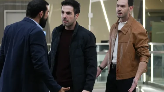 Three men in a modern, glass-walled lobby argue or confront each other; one in a brown leather jacket, another in a dark puffer, the third in a navy suit.