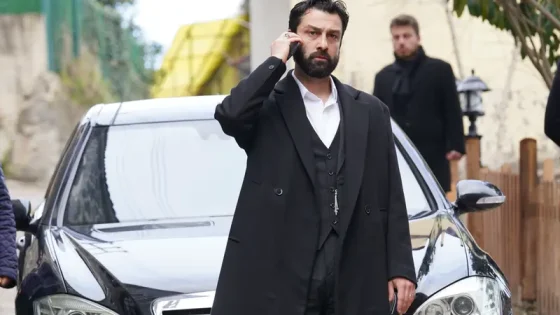 Man in a black suit and vest on a cellphone stands beside a parked dark sedan, outdoors on a sunny day.