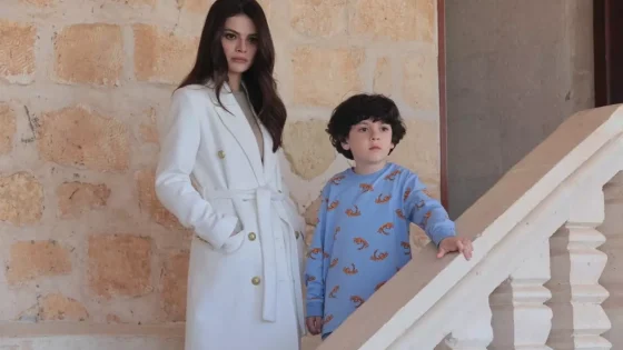Woman in a white belted coat stands beside a young boy in a blue pajama top with brown print on a stone wall and stair railing backdrop.