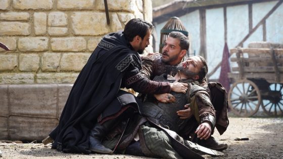 A cloaked man restrains an armored fighter while another man lies on the ground in a dusty medieval street scene