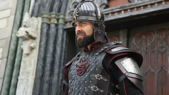 Bearded man in ornate black and silver medieval armor, helmet with star crest, standing outside a fortress doorway and looking to the side.