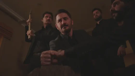 Dimly lit room; four men in black suits loom over a seated man, the foreground man looking tense at the camera.