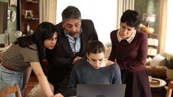 Group of four adults huddled around a laptop on a dining table, focused on the screen in a living room.
