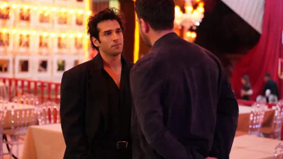 Two men in dark clothing converse in a warmly lit banquet hall with tables and chairs in the background.