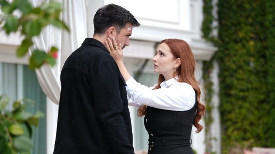 Woman with red hair cups a man's face while they exchange a tense look outside a white building; two bandages on his forehead visible.