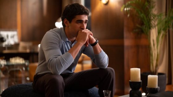 Man sits on a couch in a warm, candlelit living room, hands clasped under chin, looking thoughtful.