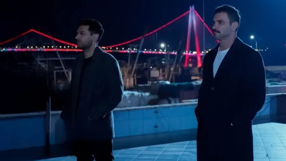 Two men stand on a waterfront promenade at night, facing left, with a red-lit suspension bridge in the background.