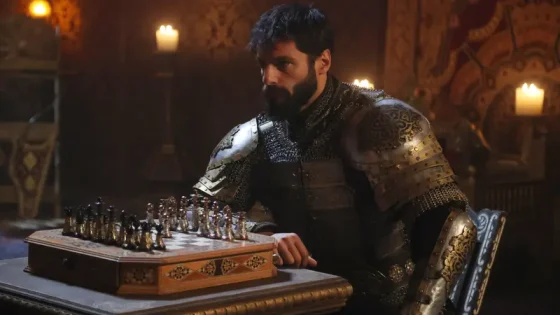 Bearded man in ornate medieval armor sits at a carved chessboard in a candlelit hall.