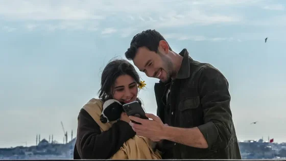 Couple standing by the water, smiling at a phone as they view it together; the woman holds a small black-and-white dog/pet in her arms.