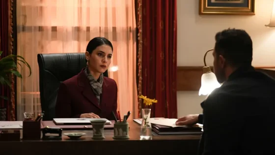 Woman in a burgundy blazer sits behind a desk, facing a man in a dimly lit office.