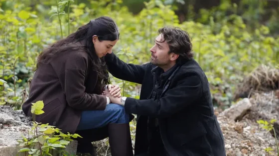 Man on one knee proposing to a woman in a leafy outdoor setting, holding her hands close.
