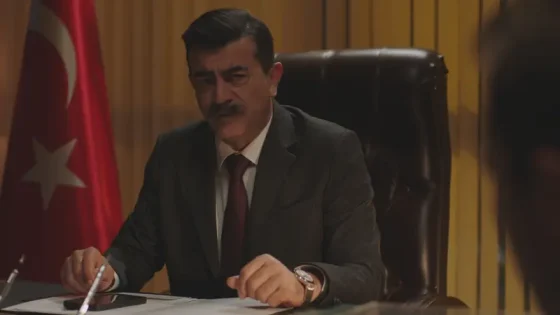 A man in a dark suit sits at a desk signing papers in an office, with a Turkish flag in the background.