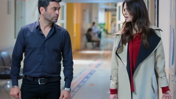 Man in a dark shirt and a woman in a beige trench coat converse in a hospital corridor.