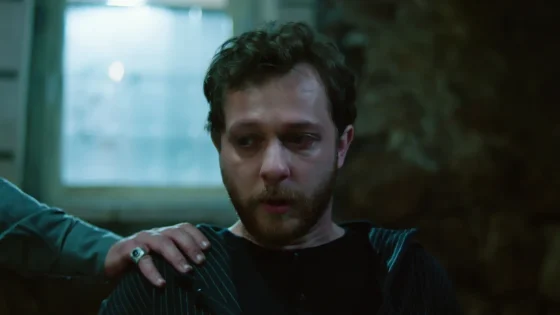 Man with a beard looks distressed as a hand grips his striped shirt on the shoulder in a dim, blue-lit room.