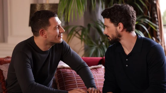 Two men sit on a red sofa facing each other and chat in a living room setting with plants in the background.