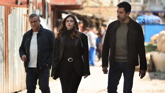 Three adults walking down a sunlit street, with a woman in the center wearing black and two men in jackets flanking her.
