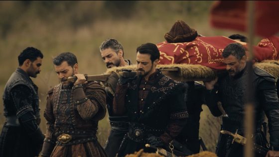Group of armored men carrying a coffin draped in red and gold cloth on a wooden bier, walking through a field.
