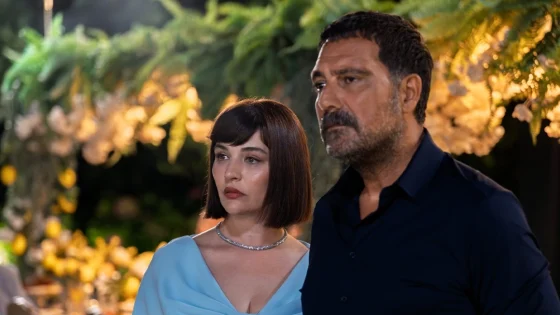 Man and woman stand together at an outdoor evening event, both with serious expressions, surrounded by warm floral lighting.