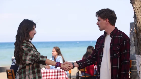 Two young adults shake hands at a beachside cafe, with the ocean in the background.