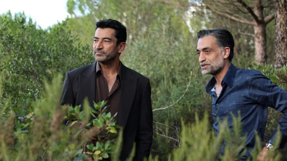 Two men stand among bushes in a lush outdoor setting, one in a dark suit and the other in a blue shirt, looking off to the side.