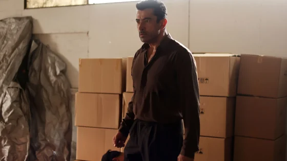 Man wearing a dark shirt standing in a storage warehouse with stacked cardboard boxes and bags around him, looking to the side.