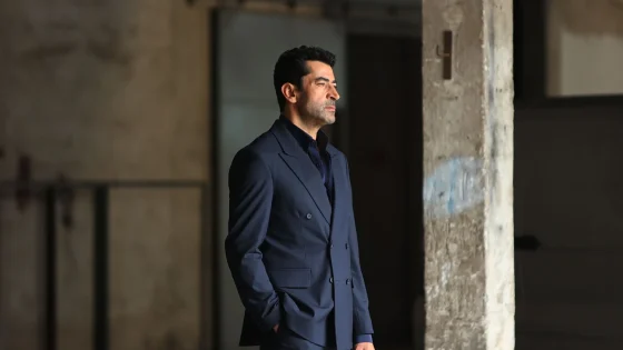 Man in a navy suit standing in a dim, industrial space, looking to the right with hands in pockets.