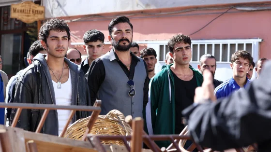 Group of men outdoors in a street scene, central man wearing a gray vest, with others around and baskets in the foreground.
