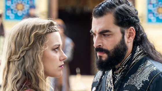 Close-up of a woman with braided blonde hair and a bearded man facing each other outdoors.