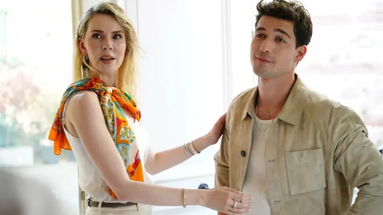 Two adults stand indoors, the woman touching the man's beige jacket as they converse.