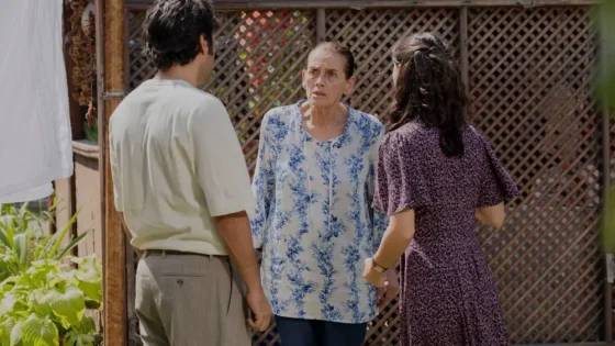 Three adults stand in a sunny courtyard, with the middle-aged woman looking surprised as the man and other woman face her.