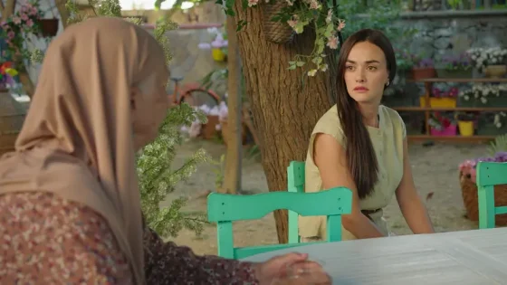 Two women sit at an outdoor table under a tree; one wears a tan hijab, the other has long dark hair and a beige top, both seated on turquoise chairs.
