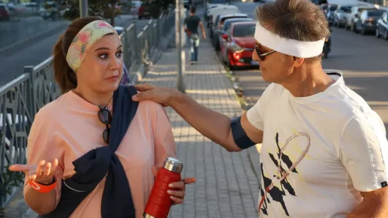 Man with a white headband consoles a woman in a pink top on a city sidewalk; he rests a hand on her shoulder while she holds a red water bottle.
