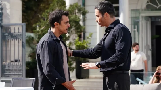 Two men stand face to face outside a building, one placing a reassuring hand on the other's chest as they talk seriously.
