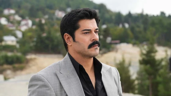Man in a light gray suit jacket and black shirt gazing to the right, outdoor hillside setting blurred in the background.
