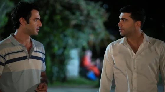 Two men stand and talk on a nighttime outdoor path with greenery in the background.