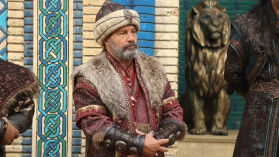 Man in ornate fur-trimmed robe and turban standing with hands clasped, in front of a patterned wall and a lion statue.