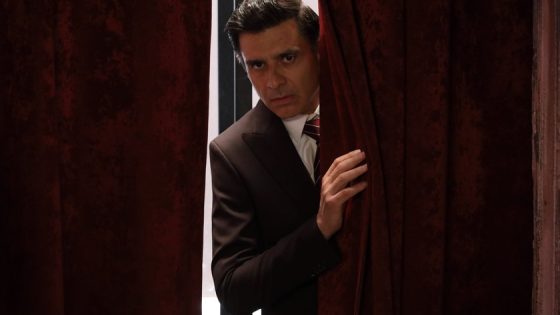 Man in a dark suit peeking from behind red velvet curtains with a serious expression.