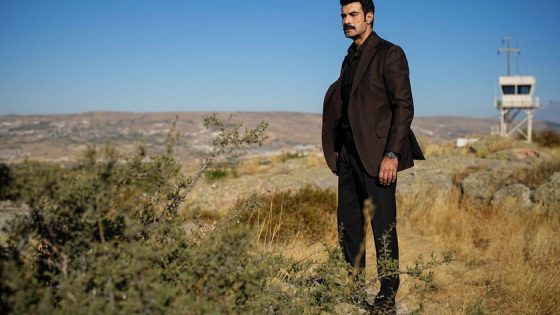Man in a dark brown blazer and black pants stands in dry scrubland with a watchtower in the distance.