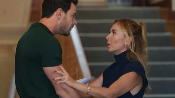 Woman with blonde hair holds a man's arm, both facing each other with tense expressions on a stairway.