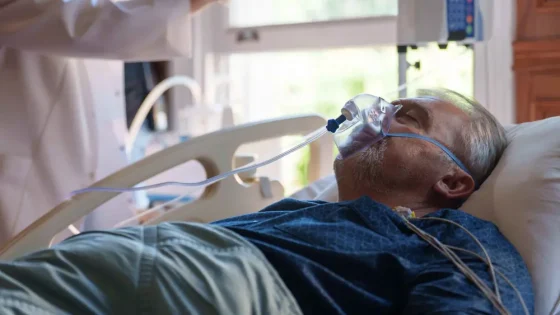 Older man lying in a hospital bed wearing an oxygen mask connected to tubing and monitors nearby.