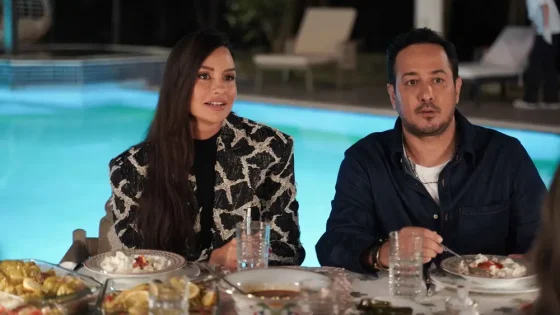 Couple dining outdoors by a lit pool at night, with plates of food and drinks in front of them; woman in a patterned jacket, man in a dark shirt.
