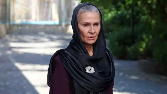 Older woman wearing a black headscarf with a silver brooch, standing outdoors on a sunlit street.
